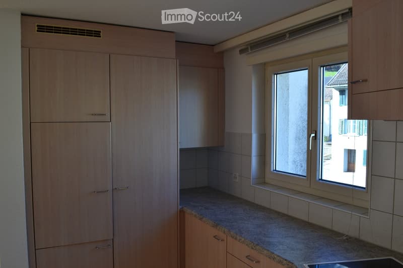 Modern kitchen with light wooden cabinets, granite countertop, and large windows offering a view of the exterior.