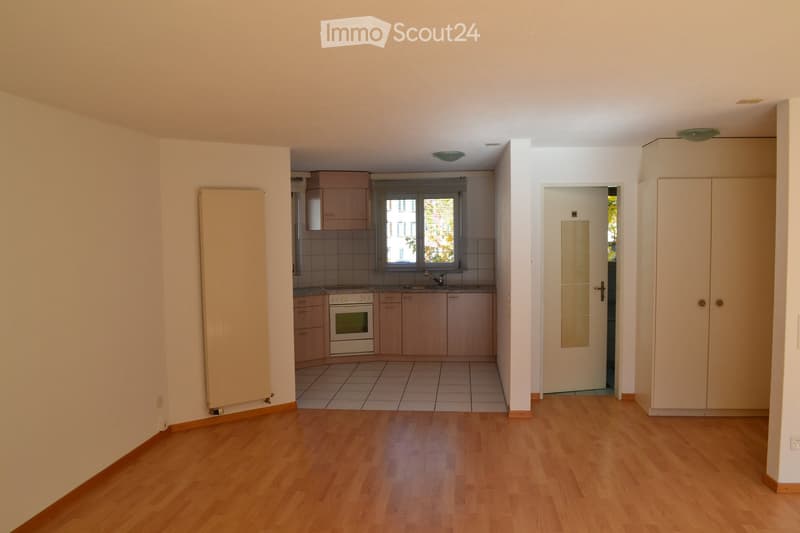 Empty room with wooden flooring, light-colored walls, and a doorway leading to a kitchen area with cabinets, a stove, and a sink.