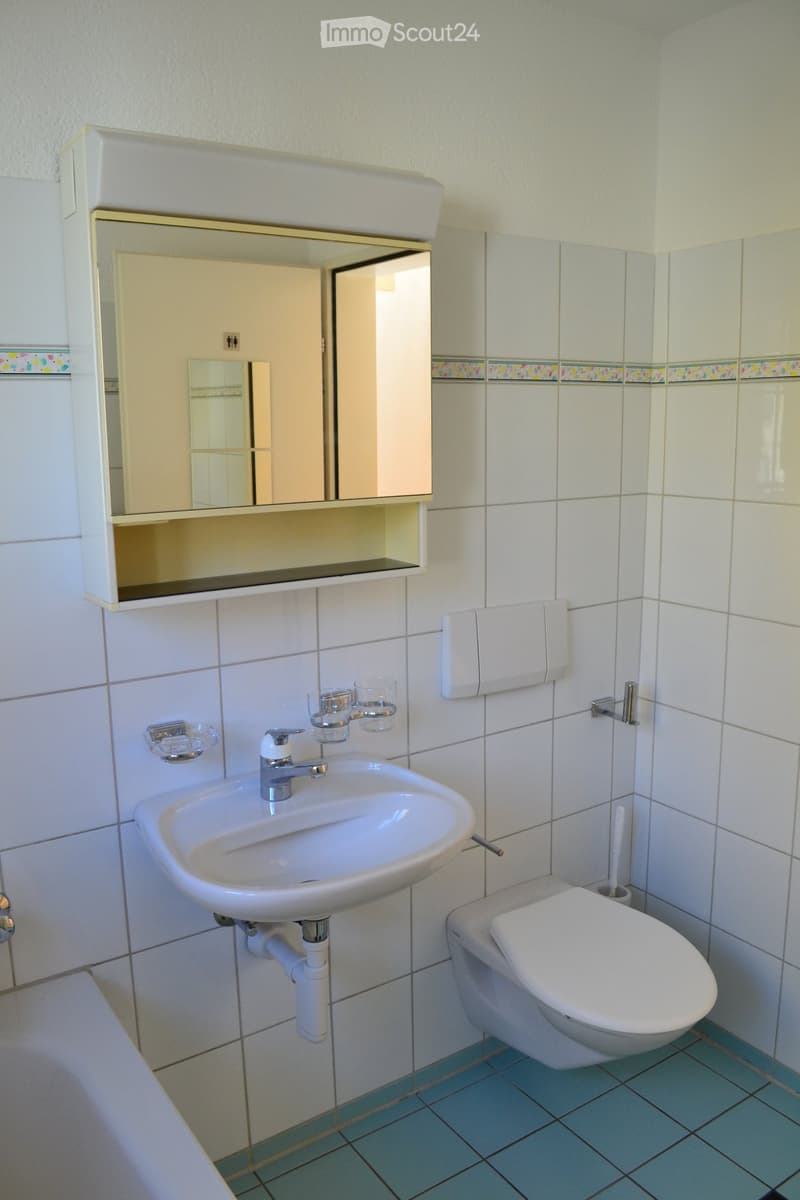 A small bathroom with a white sink, chrome faucet, mirror cabinet, toilet, and towel rack. The walls are tiled in white with a decorative border, and the floor is blue tiled.