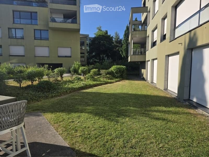 Well-maintained garden area with neatly trimmed bushes and trees, surrounded by modern apartment buildings with balconies and large windows.