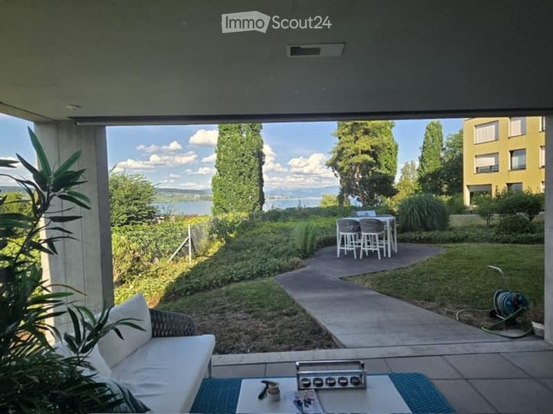 Balcony with a view of a lake, featuring a white sofa, coffee table, and a small table with chairs in the garden area.
