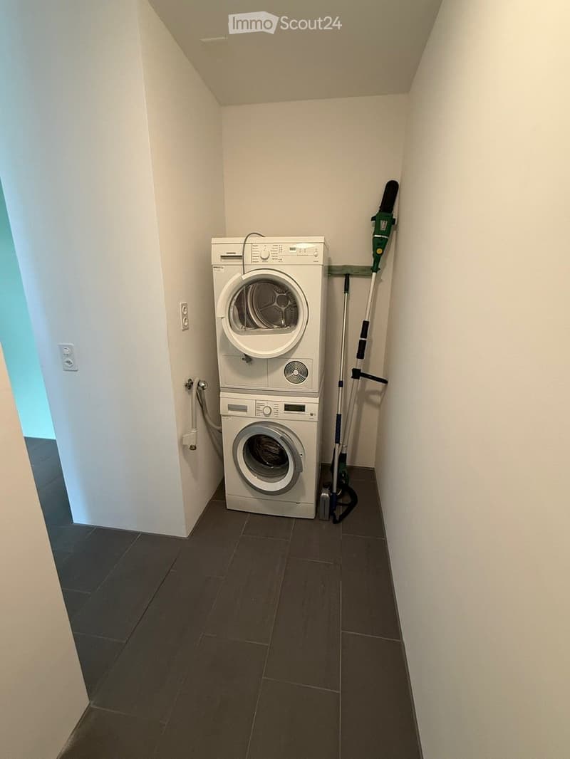 Compact bathroom with a stacked washing machine and dryer unit, dark tiled floor, white walls, and a vacuum cleaner stored on the wall.