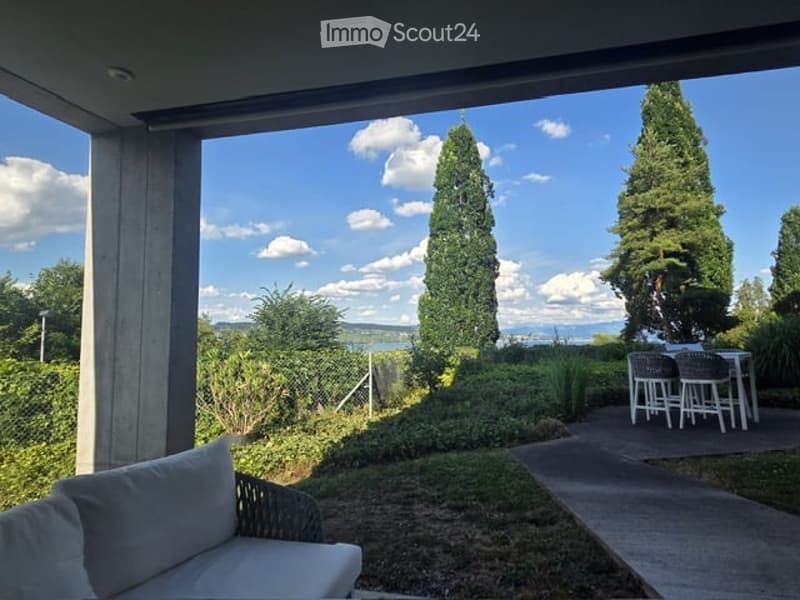 Balcony with outdoor seating area, overlooking a landscaped garden and distant view of mountains and water.