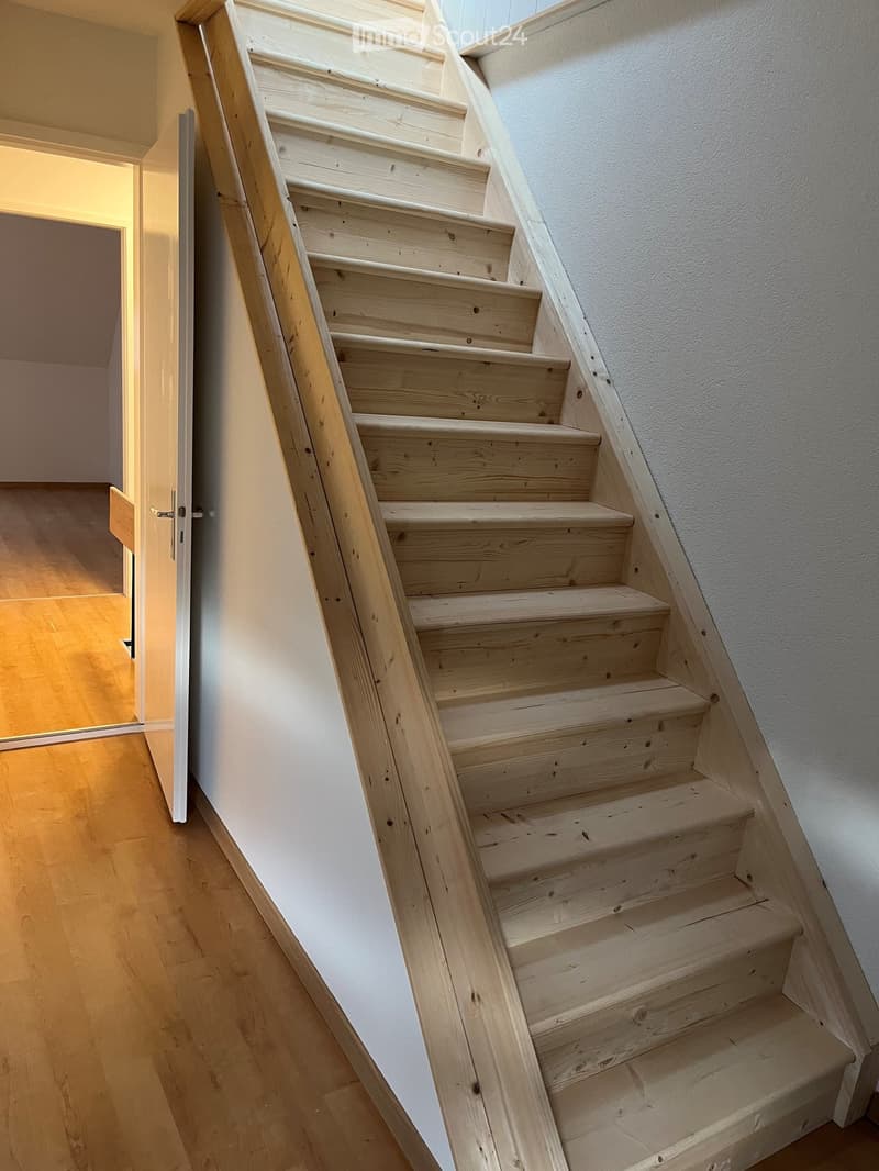 Wooden staircase with light-colored steps and railings, leading upwards, with a white wall on one side and a wooden floor on the other.