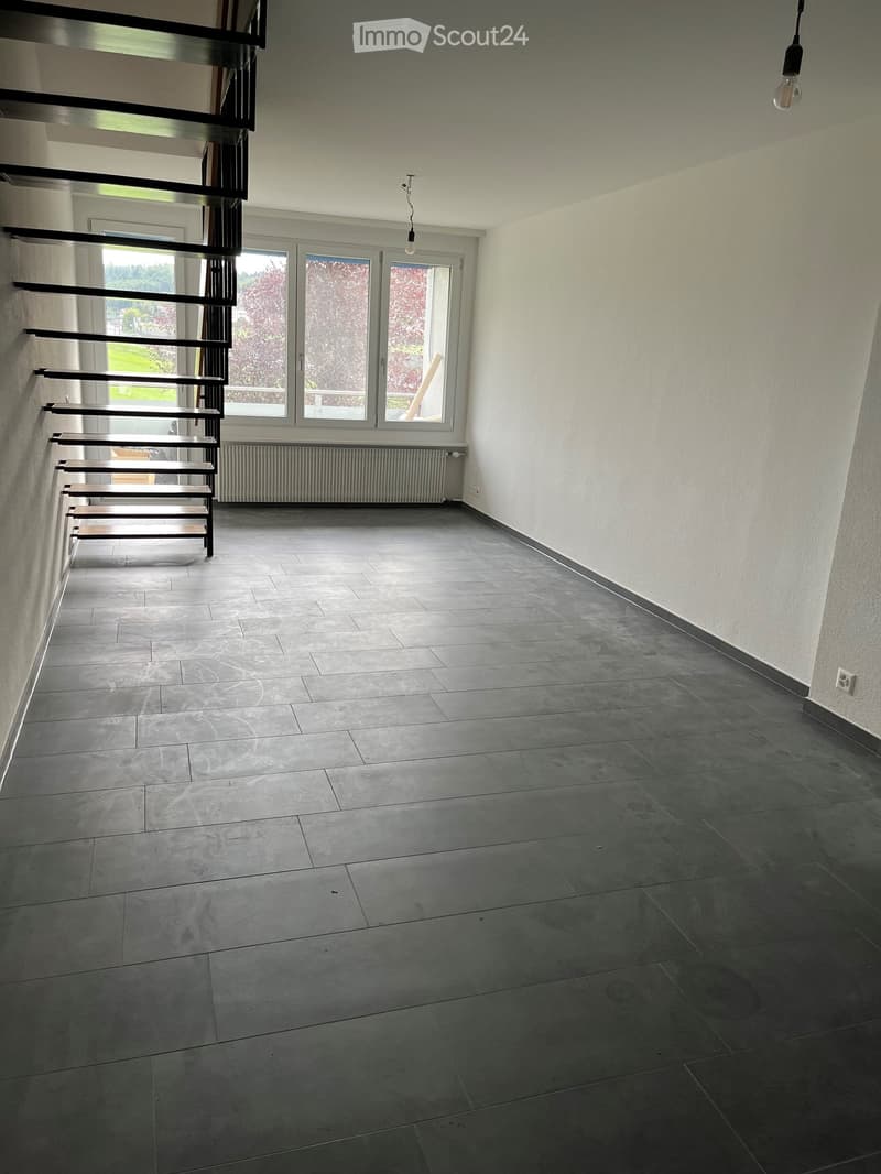 Empty hallway with dark grey tiled floor, white walls, and a staircase leading upwards. Large windows at the end provide natural light.