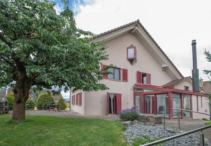 Single-story house with light pink exterior, red shutters, and a covered patio area. The house features a chimney, multiple windows, and a well-maintained garden with a large tree, lawn, and patio furniture.