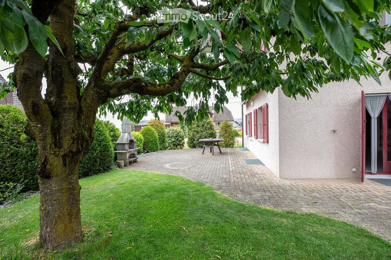 Well-maintained garden with a large tree, neatly trimmed hedges, a brick patio, a small table, and a stone barbecue area.