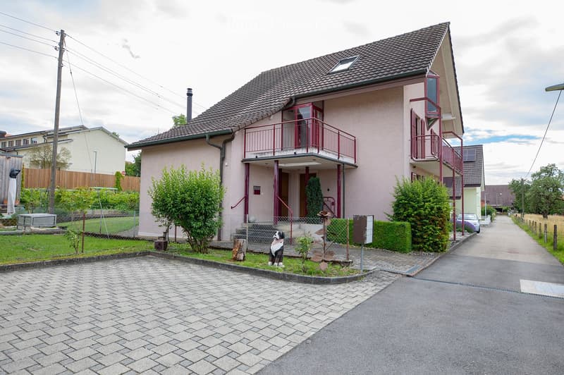 Two-story house with a tiled roof, pink facade, and red-framed balconies. The house has a small front yard with a dog statue, shrubs, and a paved driveway leading to the street.
