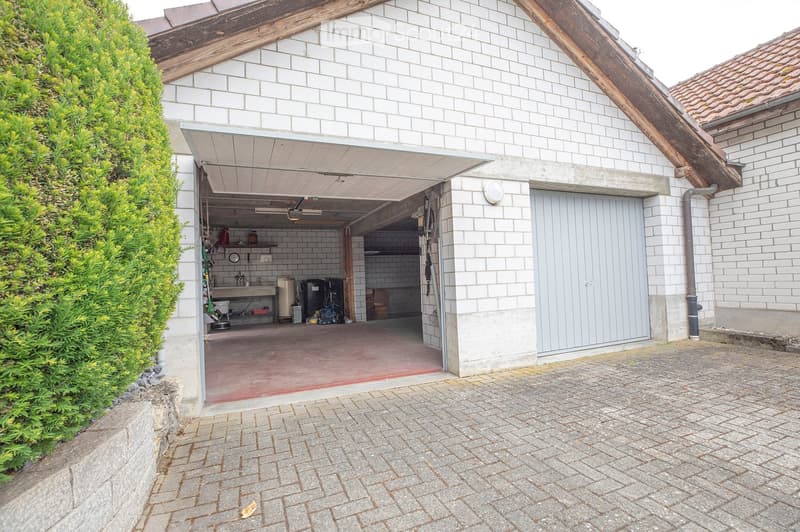 A two-story building with a large, open garage door revealing storage space inside. The garage contains shelves, tools, and various stored items. The exterior features white brick walls and a tiled roof.