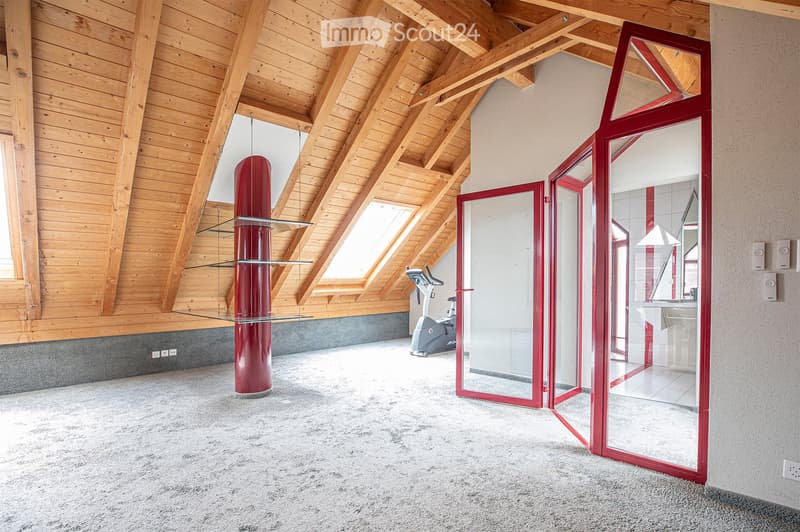 Modern, spacious room with wooden slanted ceilings, large windows, a red column, a glass table, and an exercise bike in the corner.