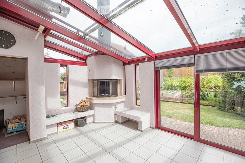 Brightly lit conservatory with a tiled floor, white walls, and a central fireplace surrounded by a bench and a wicker basket of firewood. Large windows and a glass roof allow ample natural light, with a view of a well-maintained garden outside.