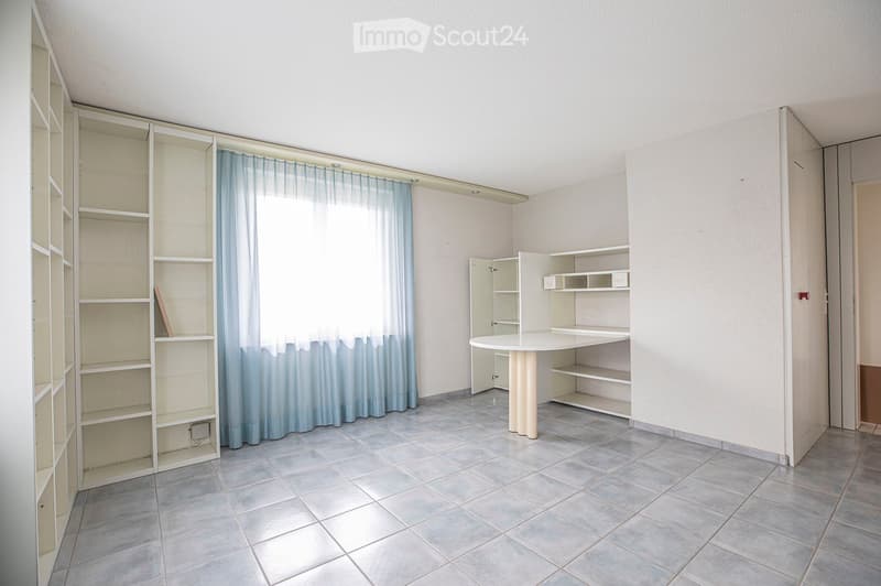 A small, unfurnished room with tiled flooring, white walls, and built-in shelves. A round table with a single leg is positioned near the window, which has a light blue curtain. The room appears to be a study or workspace area.