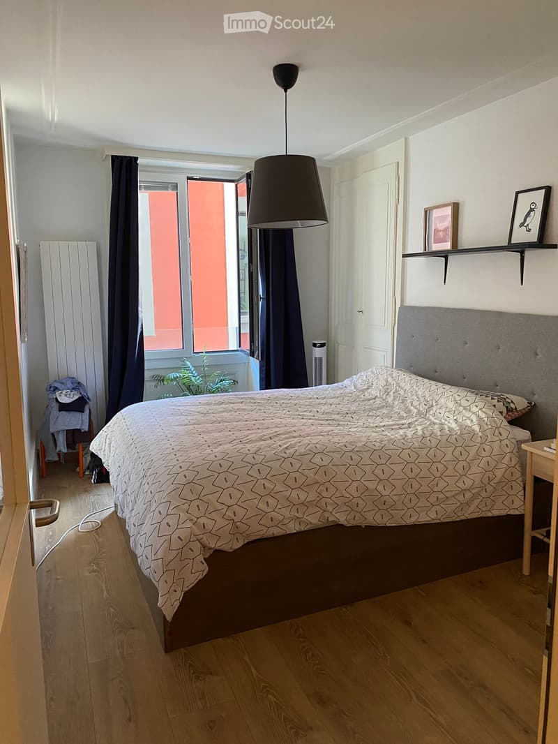 Modern bedroom with a large bed, geometric patterned duvet, pendant light, nightstand, radiator, and a view through an open door to a balcony area with plants.