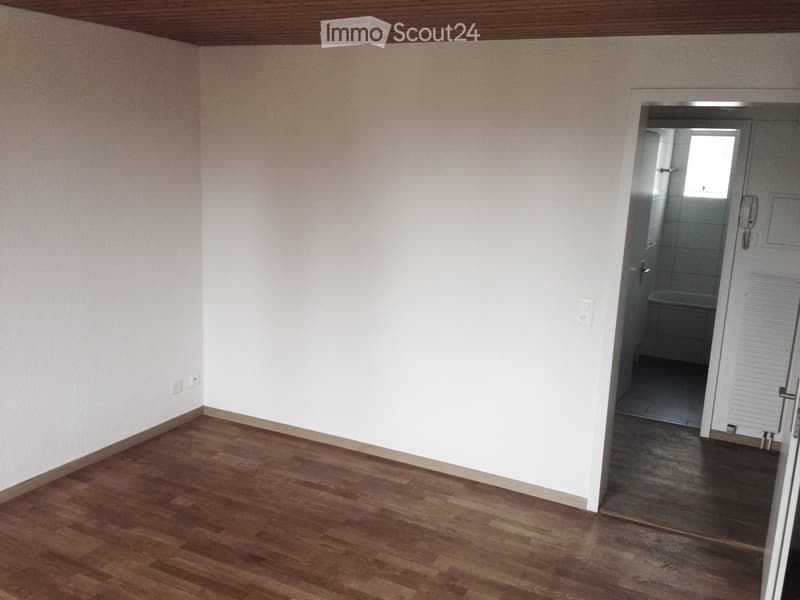 Empty room with wooden flooring and white walls, featuring a doorway leading to a small bathroom with a bathtub and tiled walls.