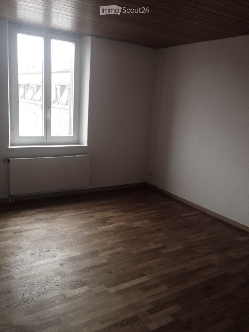 Empty room with wooden flooring, white walls, and a large window with a radiator below it.