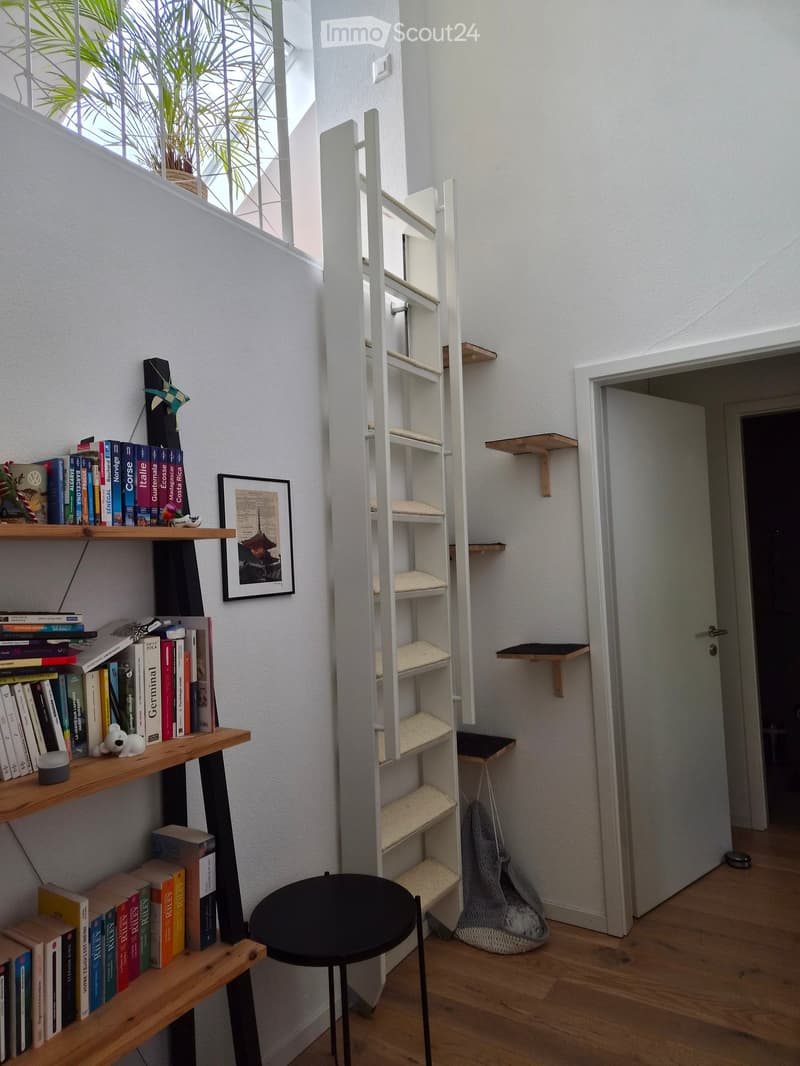 A small, modern hallway with wooden flooring, white walls, and a black stool. Shelves hold books, a cat tree, and a picture frame. A handbag is placed on the floor near the door.