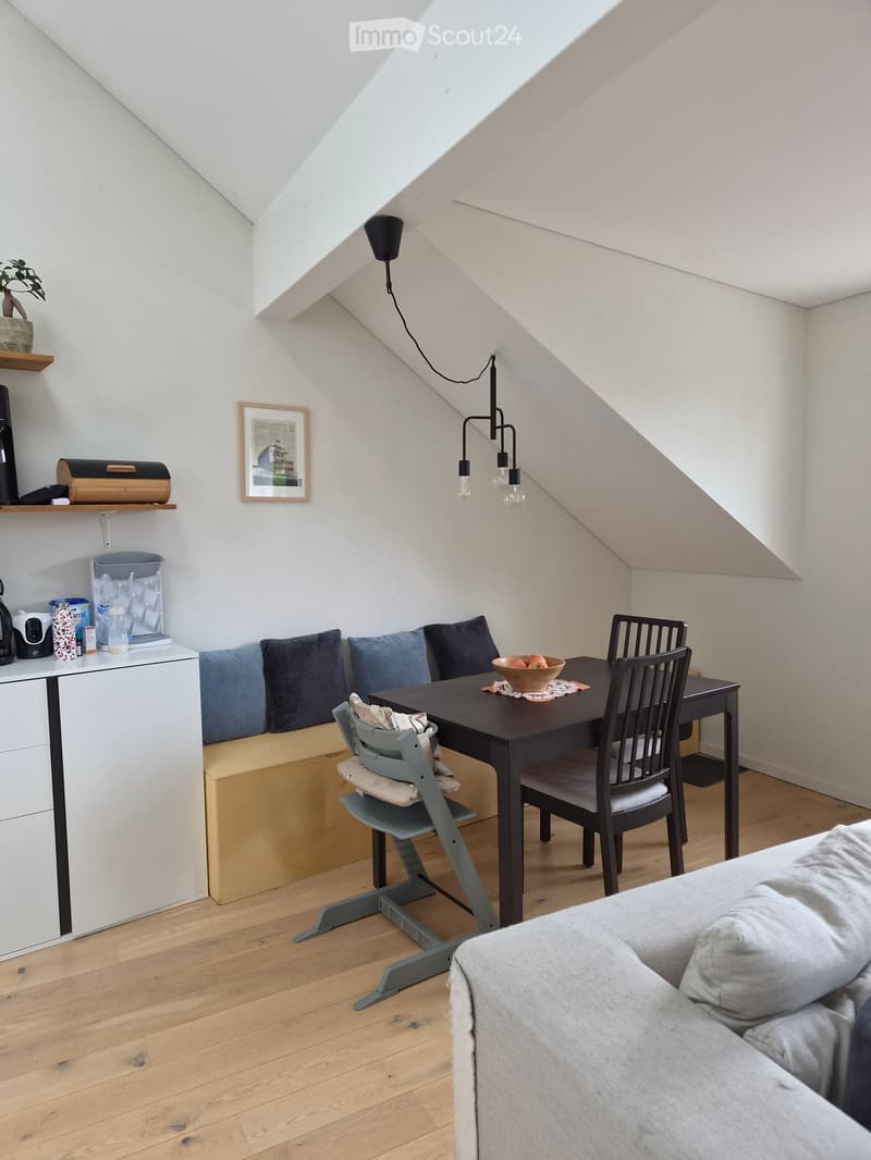 A cozy attic room with wooden flooring, a sofa, a dining table with chairs, a baby chair, a small cupboard, shelves, and a lamp.