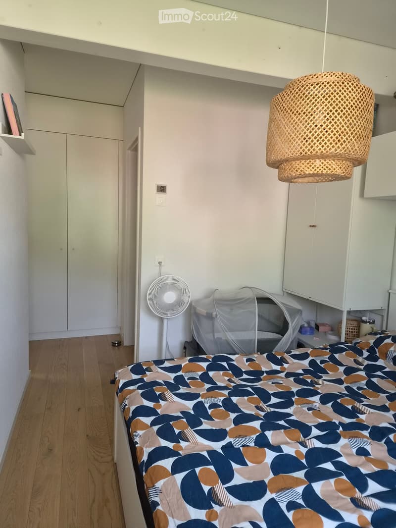 A small, modern bedroom with a bed covered in a colorful geometric-patterned duvet, a white fan, a baby crib, a wicker hanging lamp, and built-in white cabinets.
