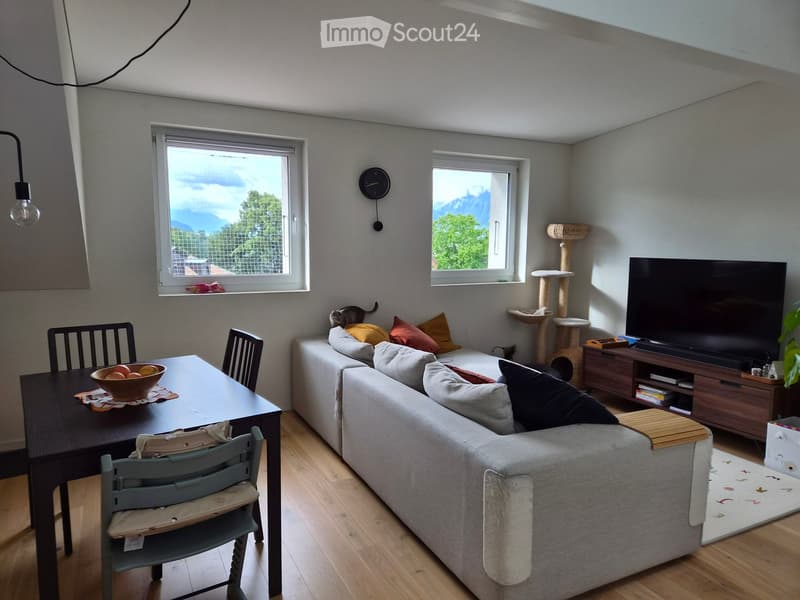 Living room with a grey sofa, wooden dining table, chairs, cat tree, television, and a bowl of fruit on the table. The room has large windows with a scenic mountain view.