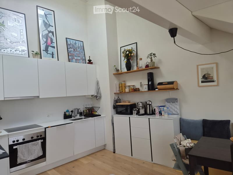 A modern, compact kitchen with white cabinets, stainless steel appliances, and a small dining area. The space features a sink, oven, microwave, coffee maker, and shelves with decorative items and kitchen essentials.