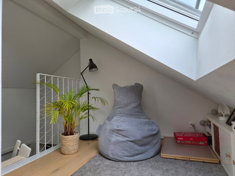 Cozy attic room with slanted ceilings, skylight, beanie chair, floor lamp, potted plant, and small shelf with books.