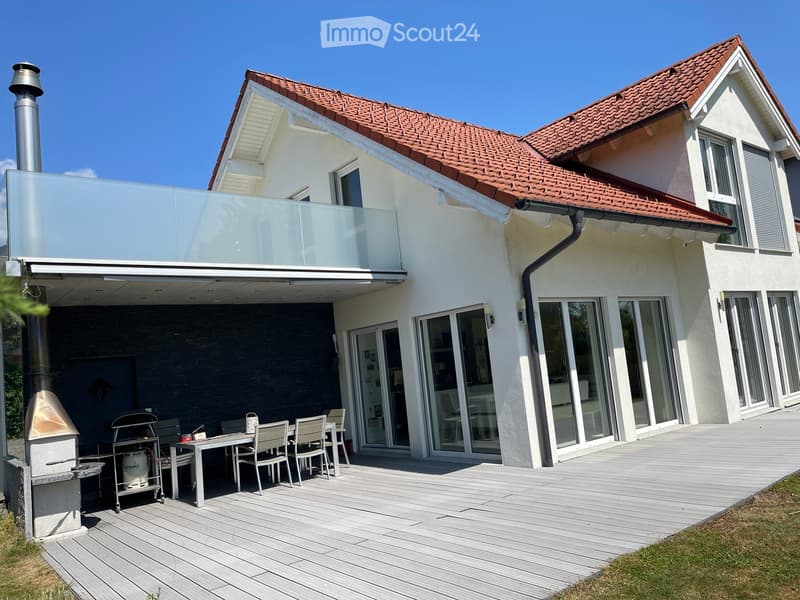 Modern two-story house with a red-tiled roof, large windows, and a spacious wooden deck with outdoor furniture including a table, chairs, and a grill.