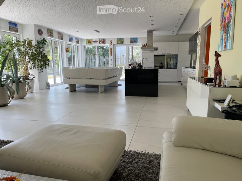 Modern living and dining area with white leather sofa, coffee table, kitchen island, and large windows. Features a sleek kitchen with white cabinets and stainless steel appliances.