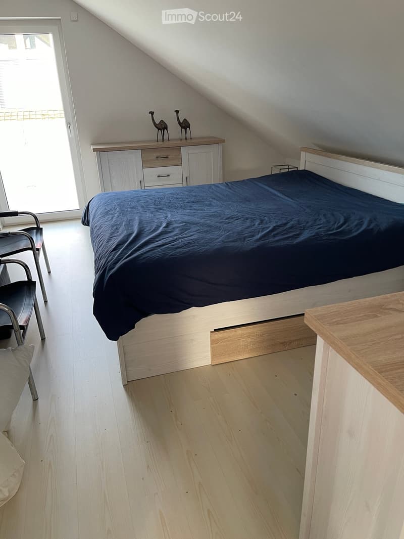 Cozy attic bedroom with a slanted ceiling, wooden floor, and large window. Features a bed with dark blue bedding, a wooden cabinet with drawers, a small table, and a chair.