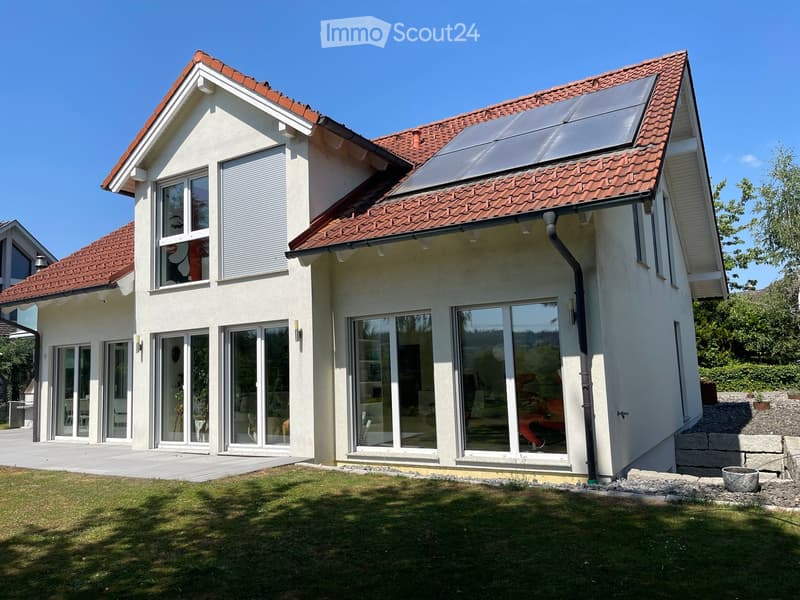 Two-story house with red-tiled roof, large windows, solar panels, and a well-maintained lawn.
