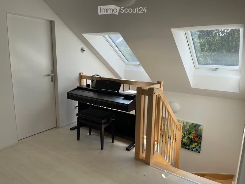 Bright attic room with wooden flooring, a black piano and stool, a staircase leading down, skylights, and a painting on the wall.