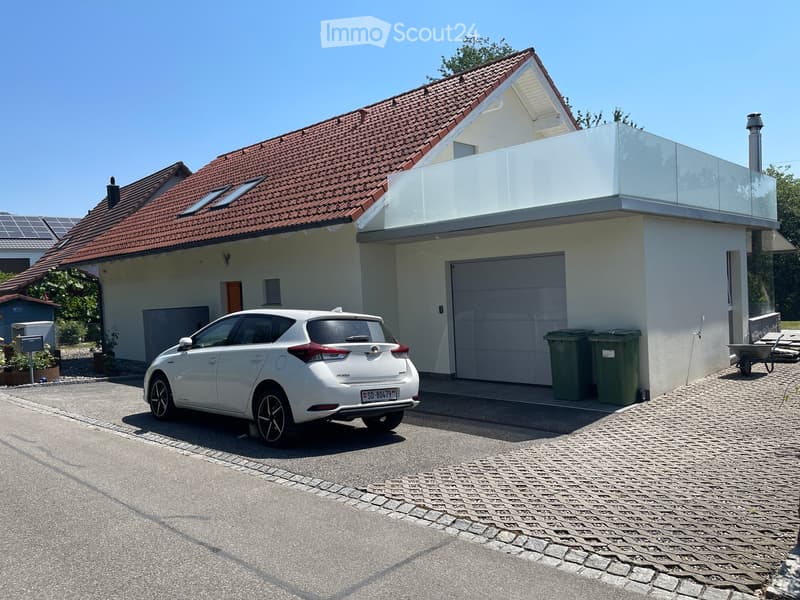 Two-story house with red-tiled roof, white walls, garage, and a modern glass balcony on the upper level. A white car is parked in front of the house.