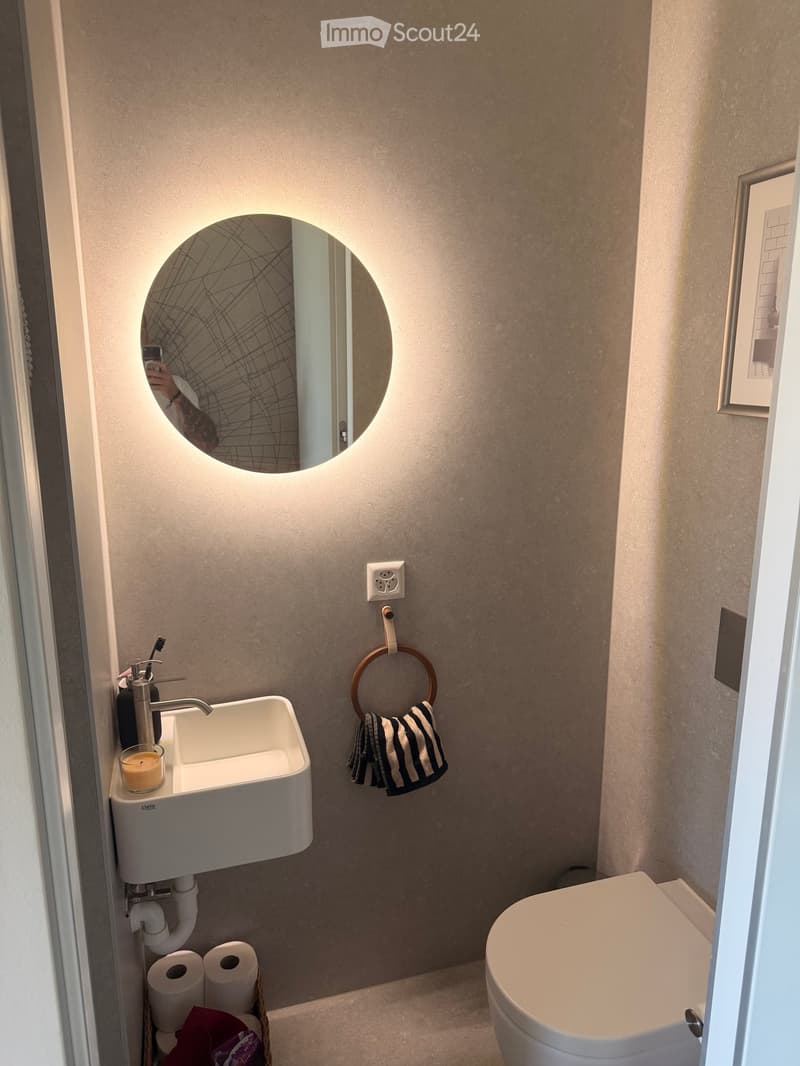 Modern bathroom with a white sink, chrome faucet, round illuminated mirror, toilet, towel ring with striped towel, and a candle on the sink.