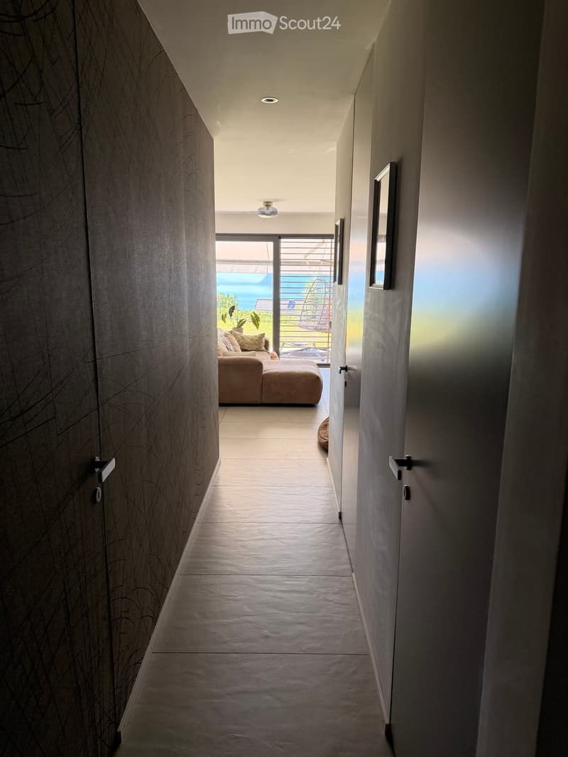 Modern hallway with sleek walls, recessed lighting, and a view of a living area with a sofa and outdoor space beyond.