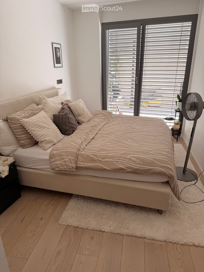 Modern bedroom with a large bed, multiple pillows, a nightstand, a fan, and a picture frame on the wall. The room features wooden flooring and large windows with blinds.