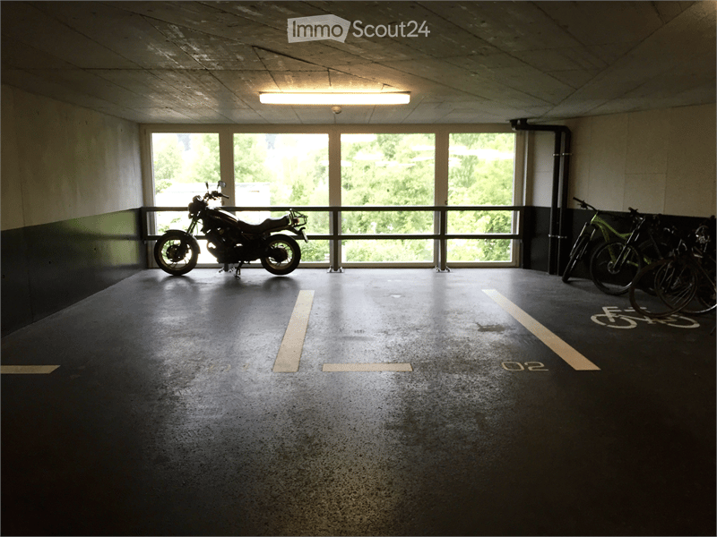 Underground parking area with a motorcycle and bicycles parked inside, large windows allowing natural light, marked parking spaces.