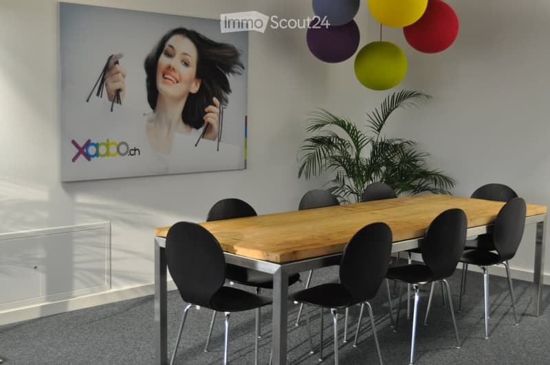 A modern meeting room with a large wooden table, black chairs, a large screen displaying an advertisement, and a potted plant in the corner.