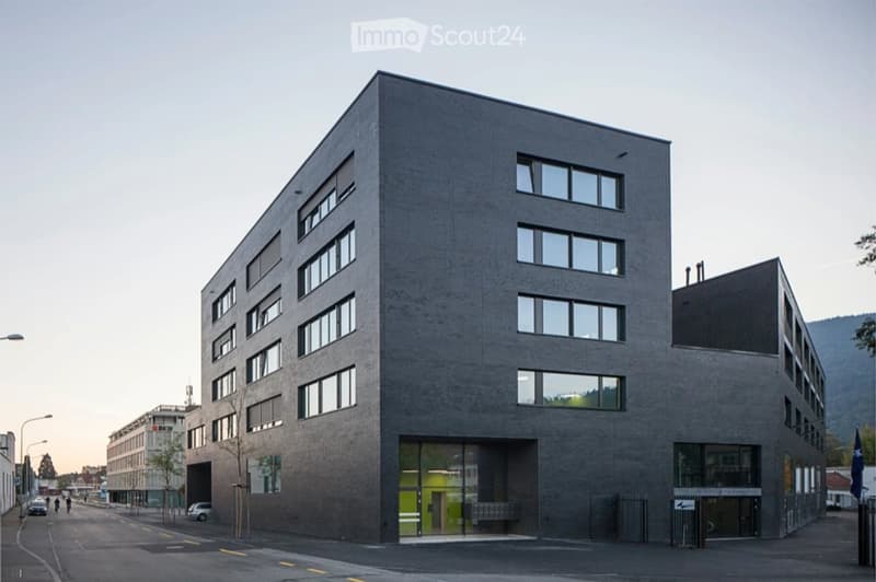Modern multi-story building with grey facade, large windows, and a glass entrance. Located on a street with a few cars and pedestrians visible in the background.
