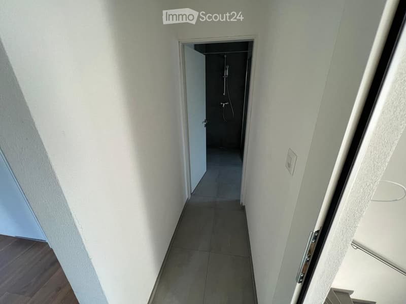 A narrow hallway with white walls and tiled floor, leading to a small shower area with a showerhead and controls.