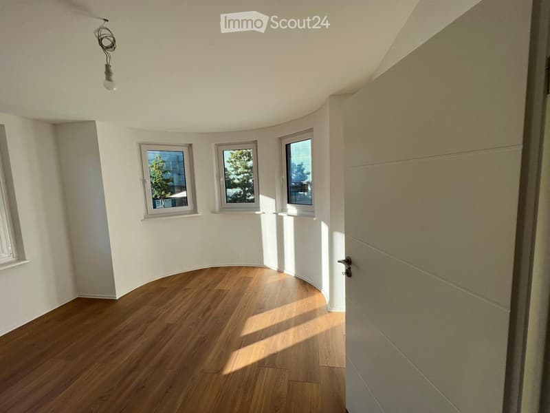 Empty room with wooden flooring, white walls, and three windows allowing natural light. The room features a curved wall design and a closed door on the right side.