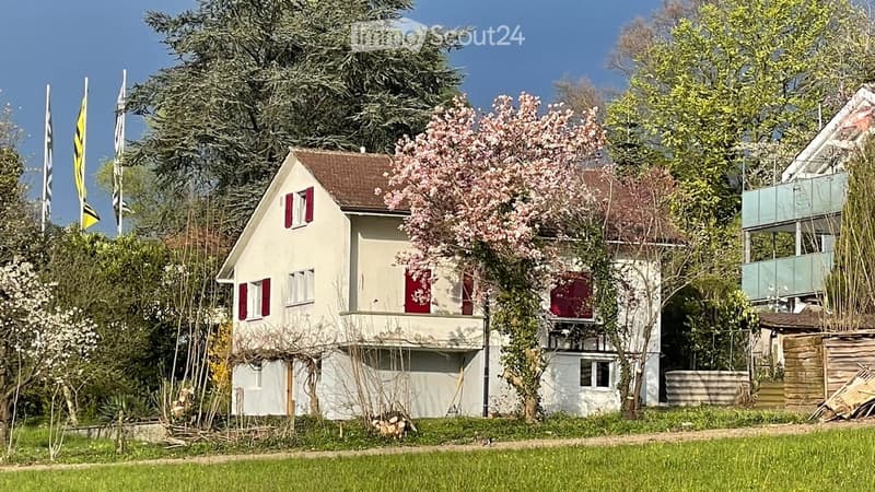 A two-story house with white walls, red shutters, and a brown roof, surrounded by trees and greenery.
