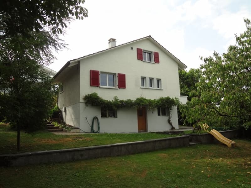 Two-story white house with red shutters, raised ground floor, small garden, and a slide in the yard.