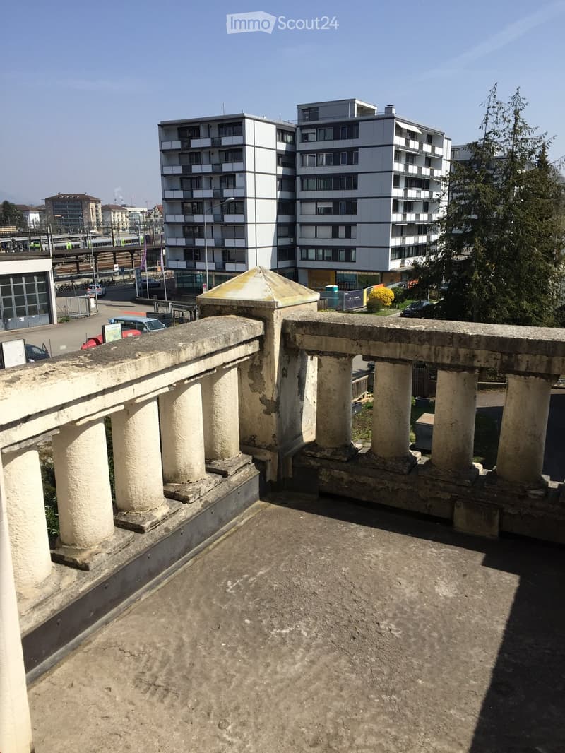 Concrete balcony with stone columns and a domed roof structure, overlooking a modern multi-story building and urban area.