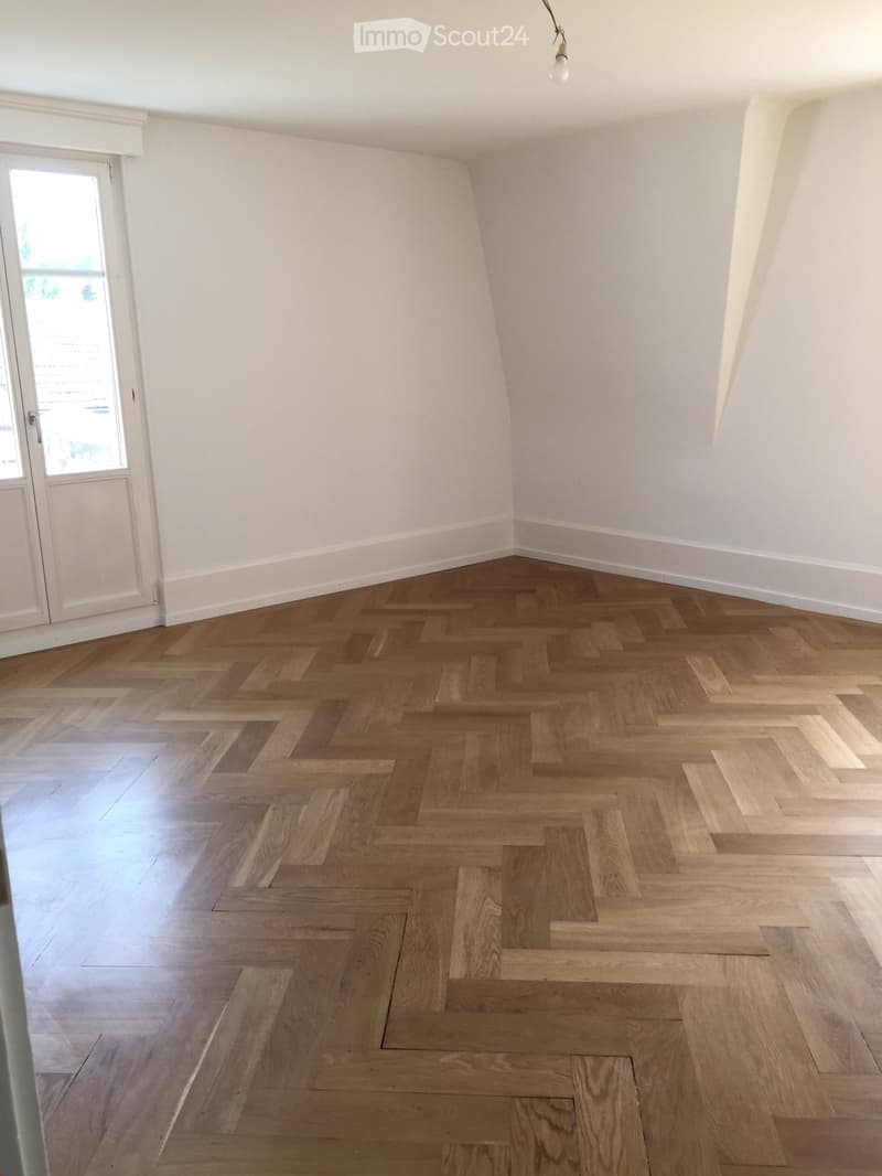 Empty room with herringbone-patterned wooden flooring, white walls, and a large window with blinds.