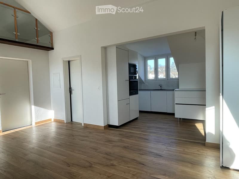 Bright, unfurnished room with wooden flooring, white walls, and large windows. Features a modern kitchen area with white cabinets and stainless steel appliances visible in the background.