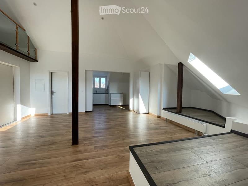 Bright, empty attic space with wooden flooring, white walls, and large windows allowing natural light. Features include exposed wooden beams, a small balcony area, and an open doorway leading to another room.