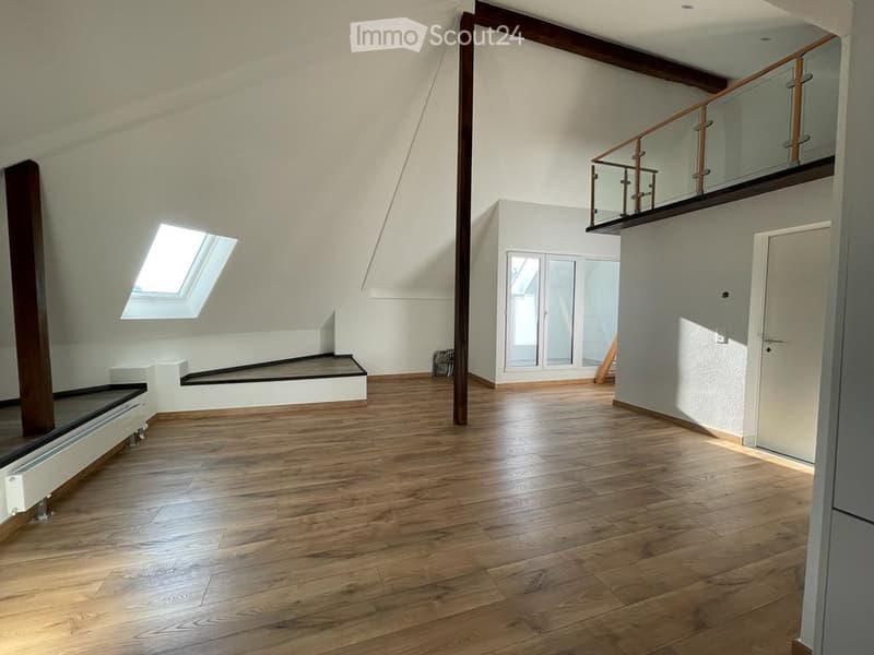 Bright, open attic space with wooden flooring, white walls, exposed wooden beams, skylight window, and a small balcony area with glass railing.