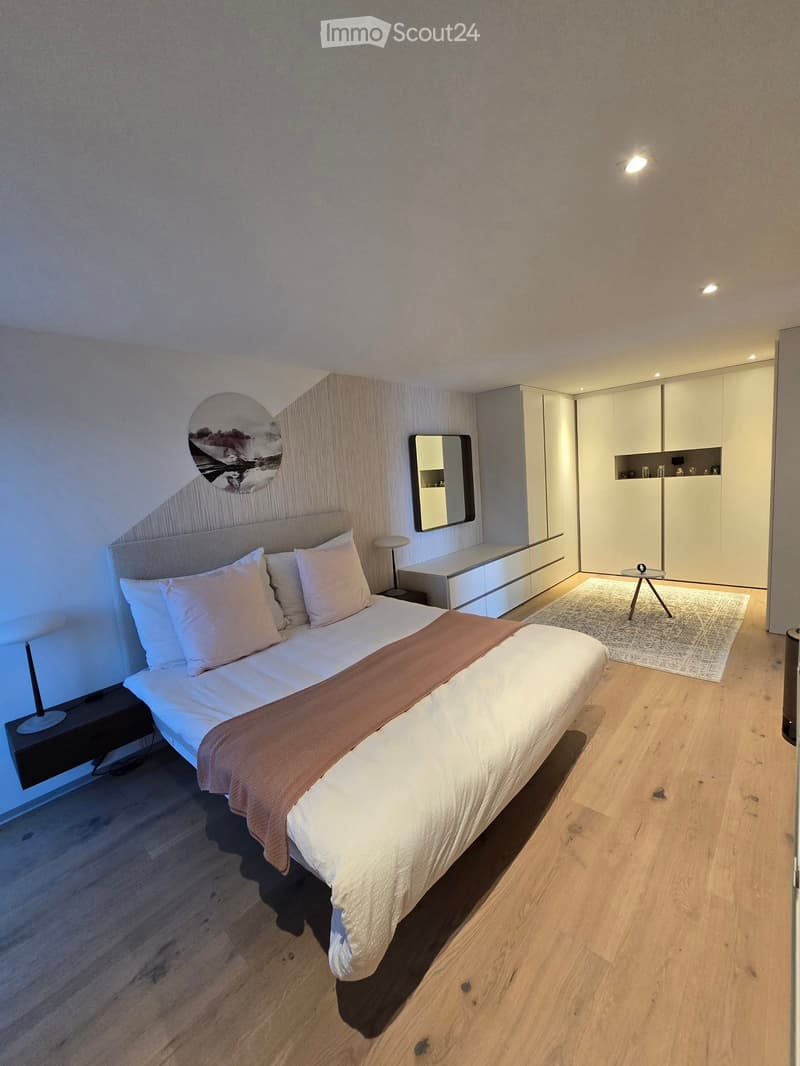 Modern bedroom with a large bed, nightstands, a wardrobe, a small table, and a rug on a wooden floor.