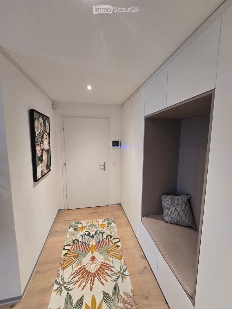 A narrow hallway with light wooden flooring, white walls, a built-in bench with a patterned cushion, a framed artwork on the left wall, and a closed door at the end.