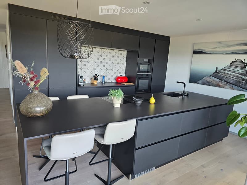 Modern kitchen with dark grey cabinets, a black island, white chairs, a vase with dried flowers, and a large painting on the wall.
