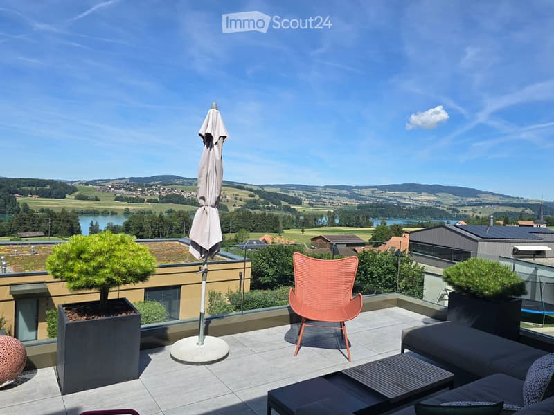 A modern rooftop terrace with a view of a lake and surrounding landscape, featuring a sofa, table, chair, umbrella, and potted plants.